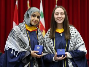 Two women in graduation gowns stand together, each holding a blue box containing a medal.