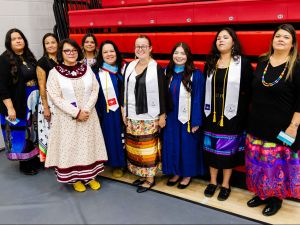 A group of Indigenous women stand shoulder to shoulder in a gymnasium smiling at the camera.