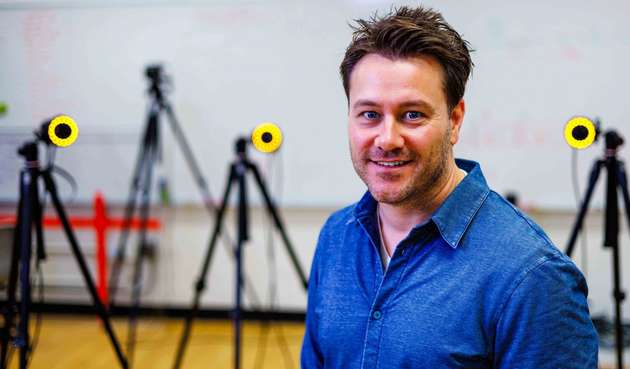 A man smiles for a photo in a lab with various motion-capture cameras in the room behind him.