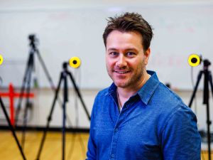 A man smiles for a photo in a lab with various motion-capture cameras in the room behind him.