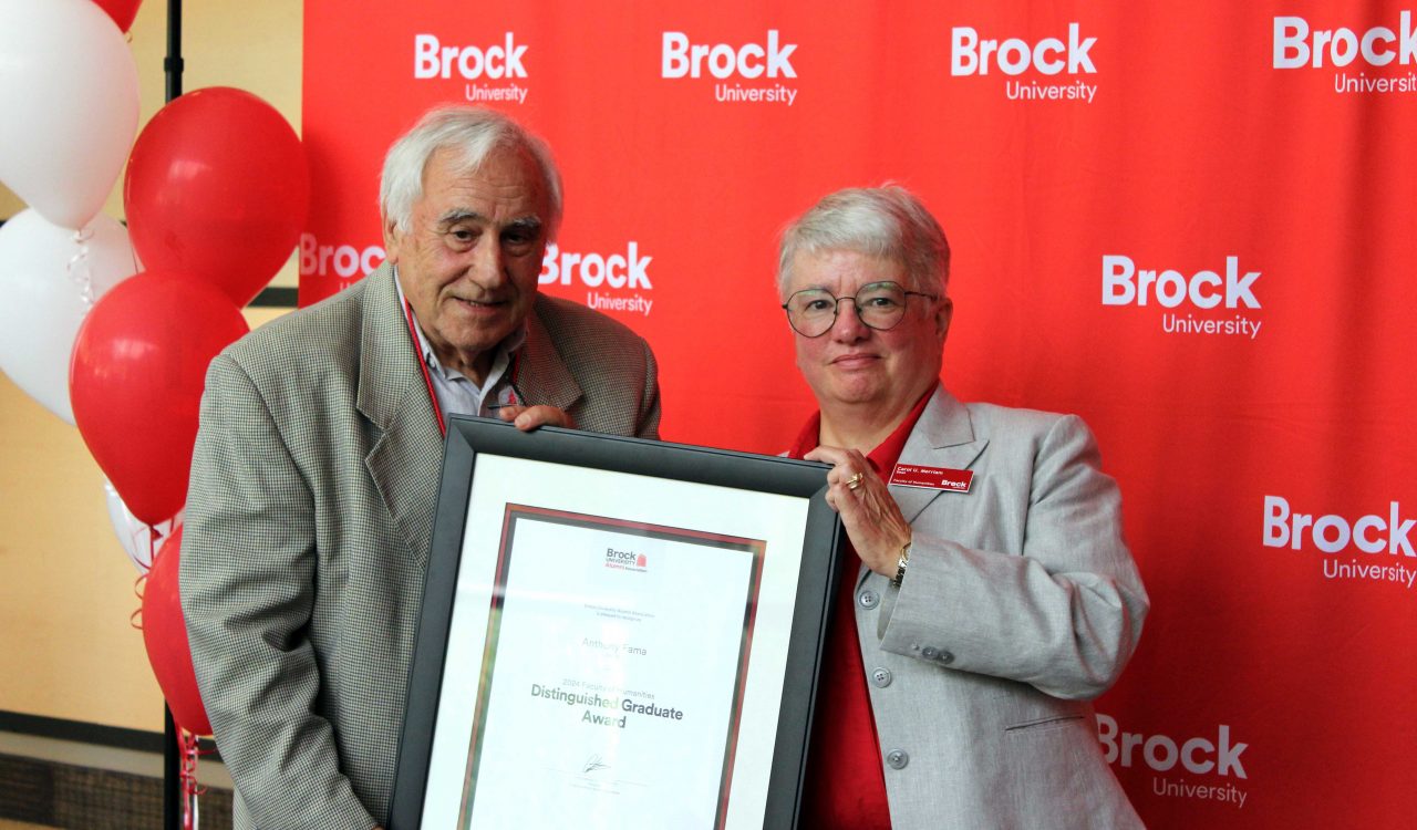 A man and woman in formal attire both hold a framed photo together smiling at the camera standing behind a bright red Brock University back drop adorned with red and white ballons.