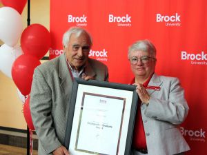 A man and woman in formal attire both hold a framed photo together smiling at the camera standing behind a bright red Brock University back drop adorned with red and white ballons.