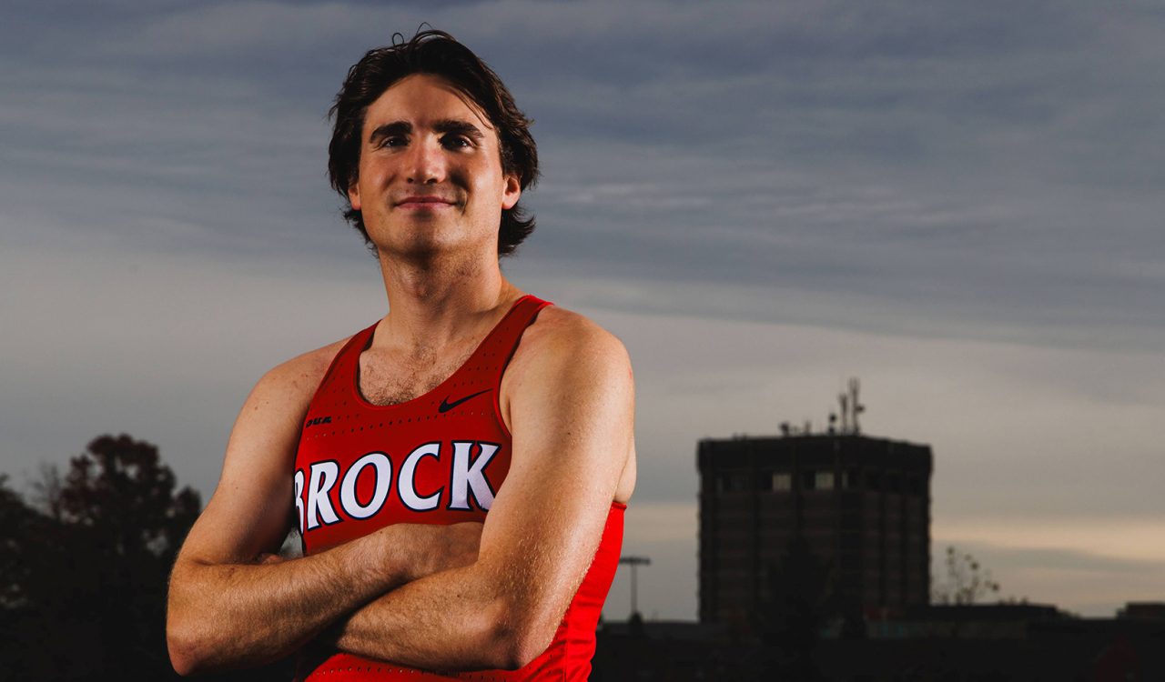 A male cross-country student-athlete from Brock University stands confidently with arms crossed, wearing a red Brock jersey. In the background, the Brock campus is visible with a tall building under a cloudy sky.