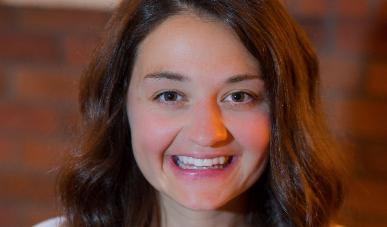 A young woman with long brown hair is shown from the shoulders up smiling warmly at the camera.