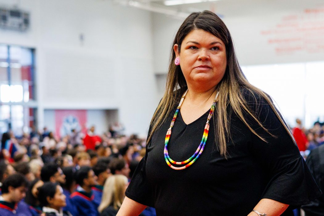 A woman with long hair and a multi-coloured necklace and dark shirt walks across the Convocation stage.