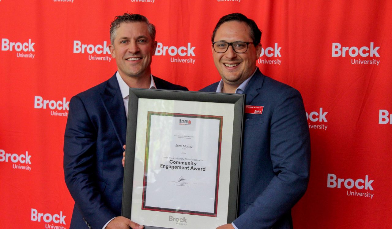 Two men pose for a photo holding a framed certificate between them.