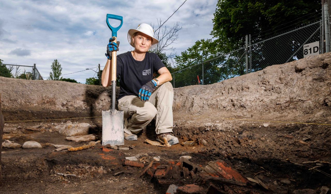 A woman holding a shovel kneels on the ground at an archeological dig site.