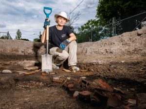 A woman holding a shovel kneels on the ground at an archeological dig site.