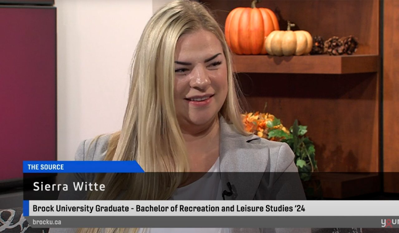 Sierra Witte wears a grey blazer and is pictured sitting in a television studio doing a media interview with pumpkins and autumn-themed decor in the background.