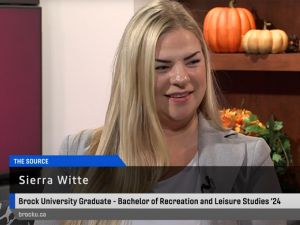 Sierra Witte wears a grey blazer and is pictured sitting in a television studio doing a media interview with pumpkins and autumn-themed decor in the background.