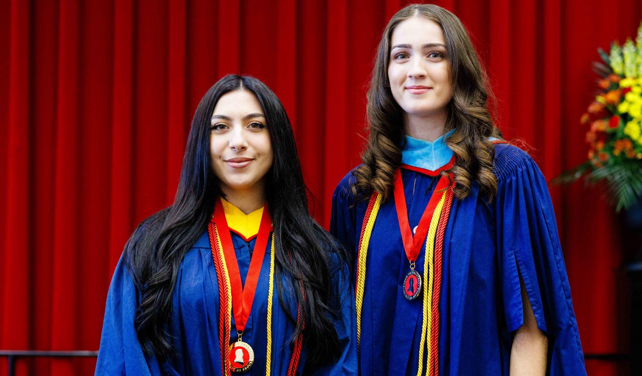 Two women in graduation gowns, each with a medal around their necks, stand together.