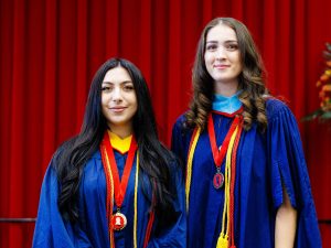 Two women in graduation gowns, each with a medal around their necks, stand together.