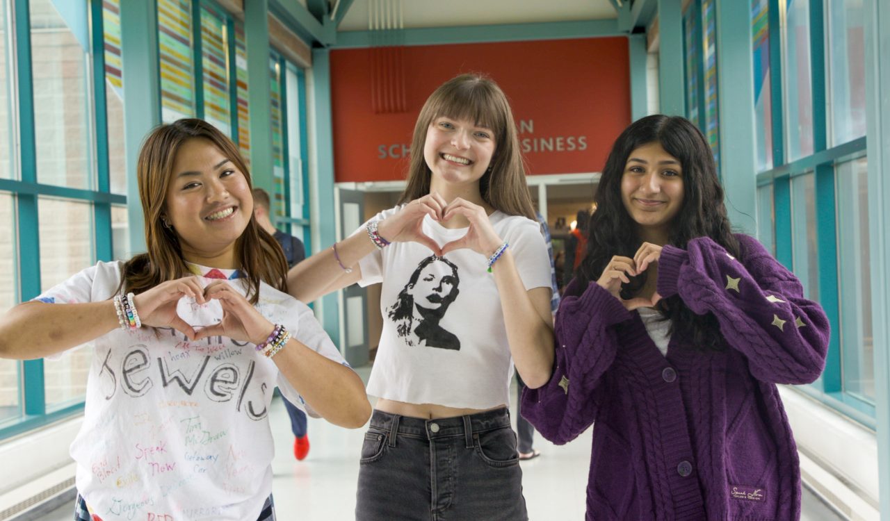 Three young women pose for a photo wearing while holding up their hands making heart gestures.