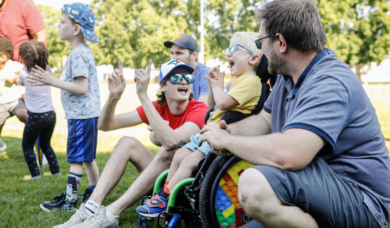 Group of children and adults enjoying outdoor activities on a sunny day. A young boy in a wheelchair, with brightly coloured wheels, laughs and claps along with others. A man beside him crouches down to chat, while a young adult in a red shirt and blue hat cheers enthusiastically. Other children stand nearby, participating in the lively moment, surrounded by green grass and trees in the background.