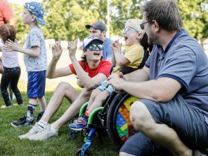 Group of children and adults enjoying outdoor activities on a sunny day. A young boy in a wheelchair, with brightly coloured wheels, laughs and claps along with others. A man beside him crouches down to chat, while a young adult in a red shirt and blue hat cheers enthusiastically. Other children stand nearby, participating in the lively moment, surrounded by green grass and trees in the background.