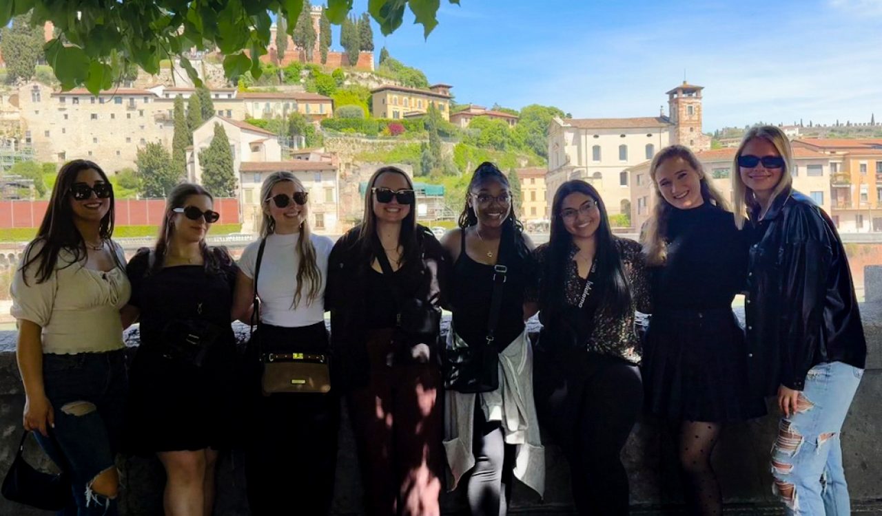 A group of female students in casual attire stand in front of a picturesque Italian village smiling at the camera on a sunny day.