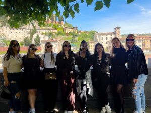 A group of female students in casual attire stand in front of a picturesque Italian village smiling at the camera on a sunny day.