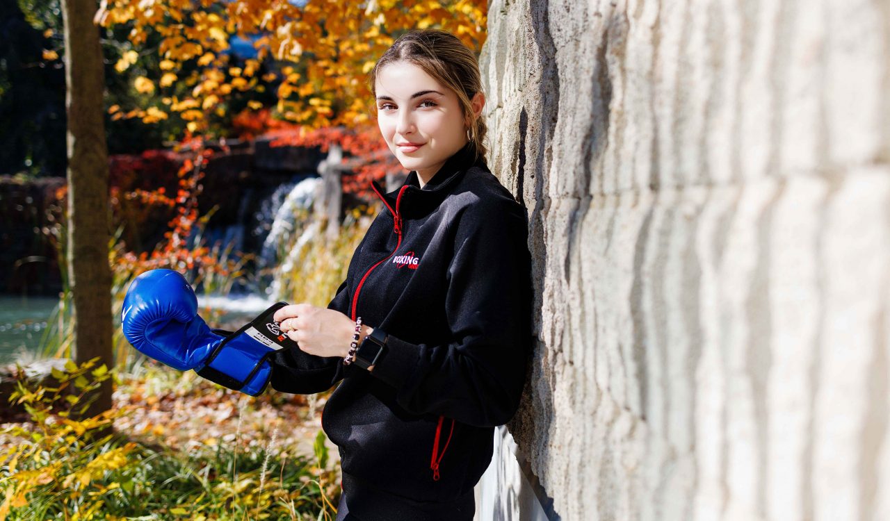 A woman leans up against a brick while equipping a blue boxing glove outside at Brock University’s Pond Inlet.