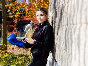 A woman leans up against a brick while equipping a blue boxing glove outside at Brock University’s Pond Inlet.