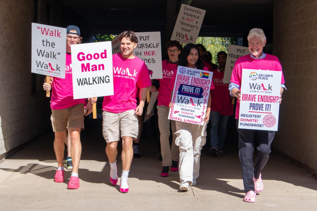 A group of people, many of them in pink shoes, walk together holding signs in support of the Walk a Mile in Her Shoes initiative in support of Gillian's Place.