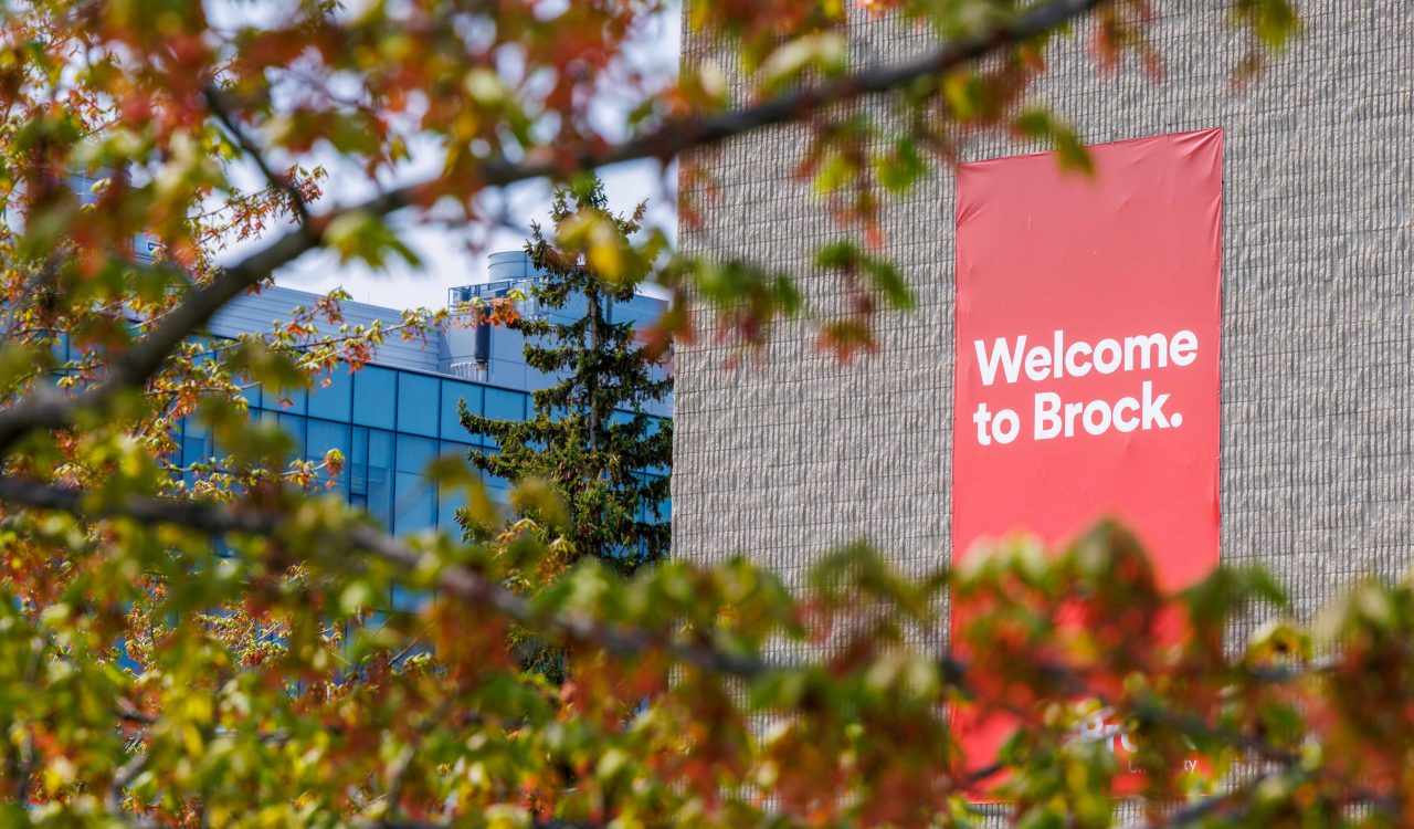 A red banner hangs outdoors on a university building with the words “Welcome to Brock” while being framed by a tree turning fall colours at Brock University’s main campus.