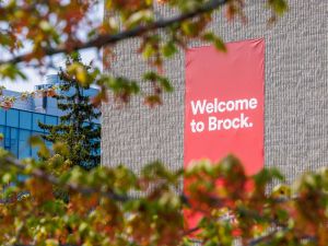 A red banner hangs outdoors on a university building with the words “Welcome to Brock” while being framed by a tree turning fall colours at Brock University’s main campus.