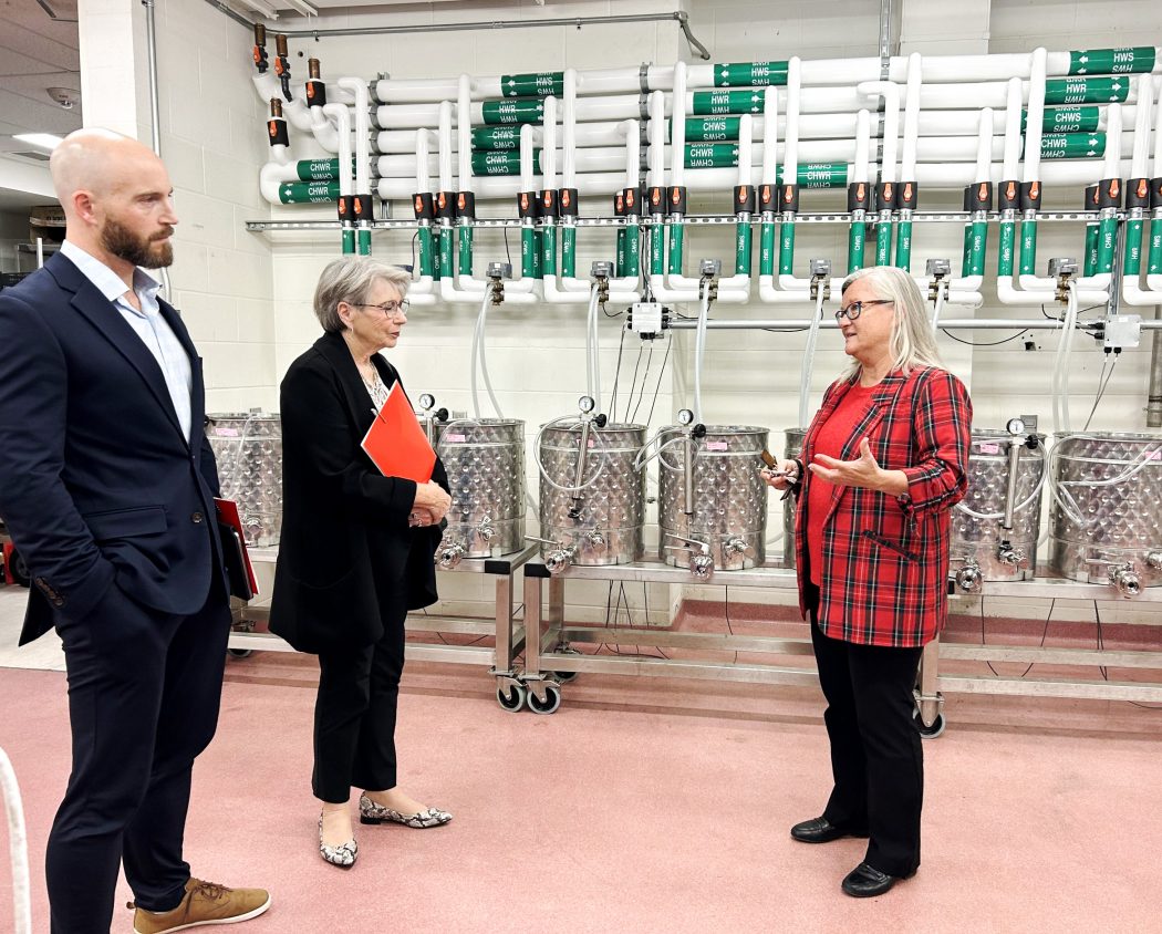 Three people stand in a room with pipes and metal cannisters on the wall in the background.