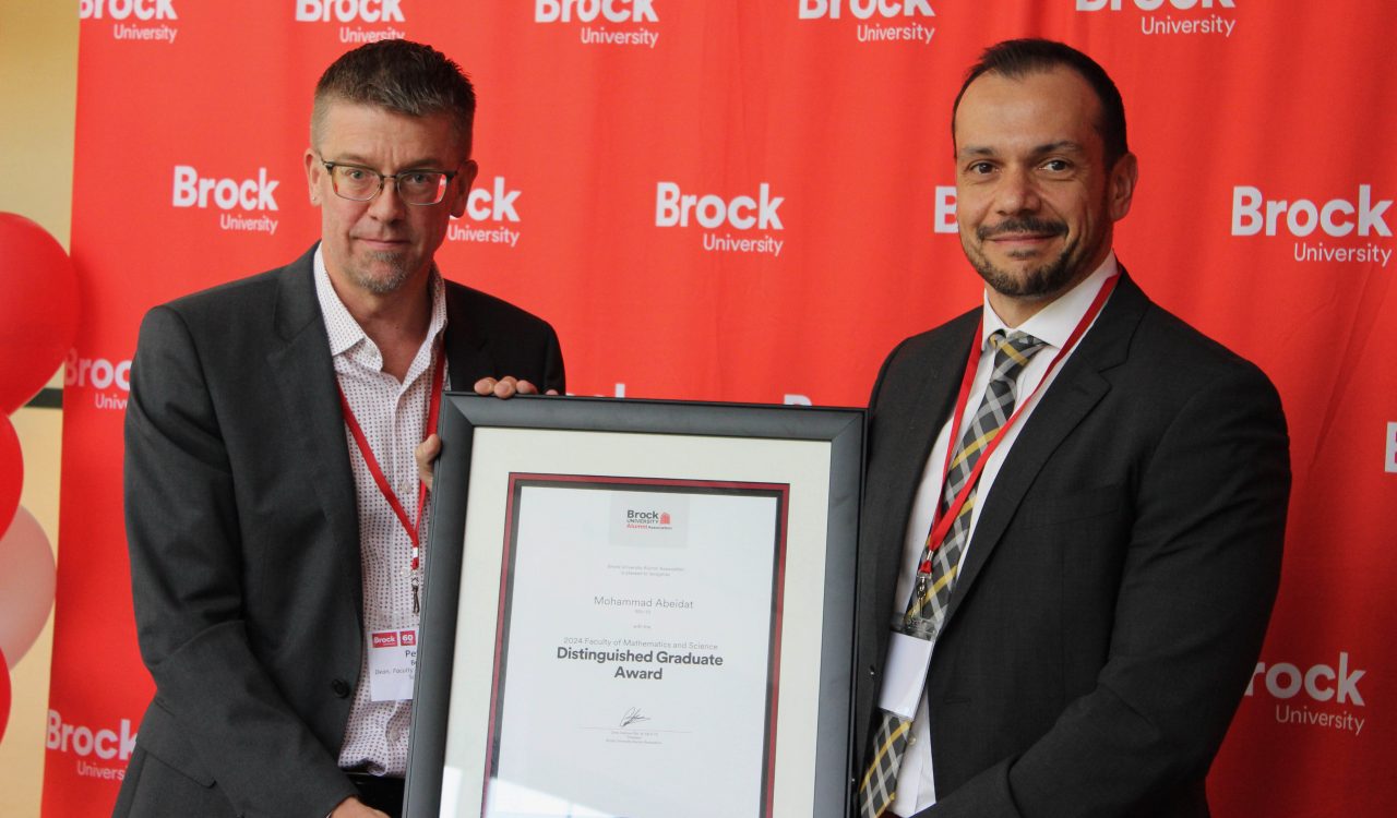 Peter Berg and Mohammad Abeidat jointly hold a framed award while standing in front of a red backdrop with many Brock University logos on it.