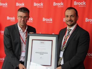 Peter Berg and Mohammad Abeidat jointly hold a framed award while standing in front of a red backdrop with many Brock University logos on it.