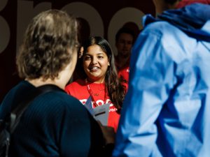 A woman wearing a red Brock University T-shirt smiles while interacting with a group of people.