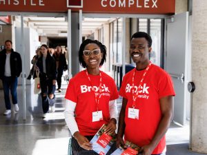 A pair of representatives of Brock University stand in a brightly-lit busy hallway smiling while wearing red Brock T-shirts and holding booklets.