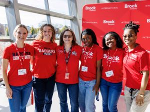 A group of six women lock arms and pose with a smile while wearing red Brock University-branded clothing against a glass wall and Brock University backdrop.