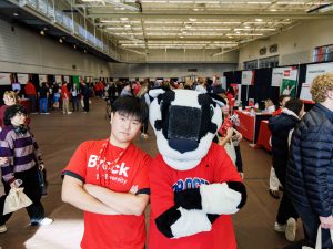 A man and Brock University’s mascot, Boomer the Badger, stand back-to-back with their arms crossed against a crowded gymnasium full of prospective students.