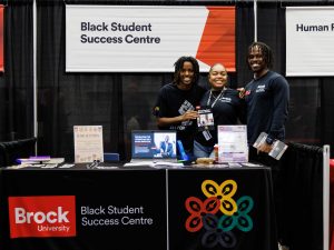 A trio of individuals stand smiling behind a Brock University booth labelled Black Student Success Centre.