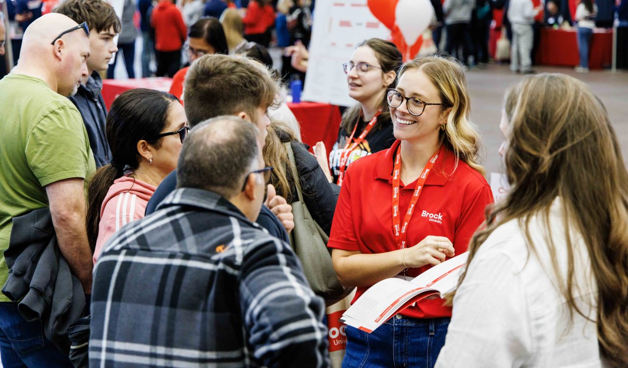 A Brock University representative wearing a red polo shirt and lanyard smiles while engaging with a group of prospective students and parents during an open house event. The room is filled with people and decorated with university-themed balloons and signs.