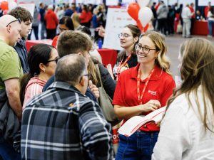 A Brock University representative wearing a red polo shirt and lanyard smiles while engaging with a group of prospective students and parents during an open house event. The room is filled with people and decorated with university-themed balloons and signs.