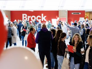 A large crowd of people tour a naturally-lit space with a large red wall with Brock University labeled in white letters serves as the backdrop. The room is decorated with university-themed balloons and signs.