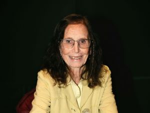 An elderly woman wears glasses and a yellow jacket, holding an award sculpture and smiling at the camera.