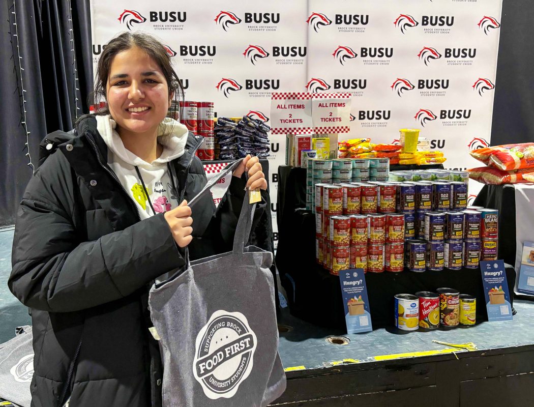 A woman holding a grey tote bag that says 'Food First' on it stands in front of a table filled with canned goods.