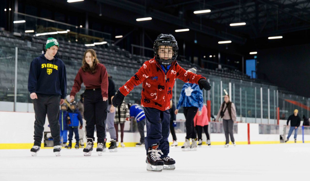 A young boy skates at an ice rink with adults and other children skating in the background.