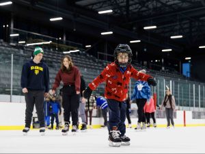 A young boy skates at an ice rink with adults and other children skating in the background.