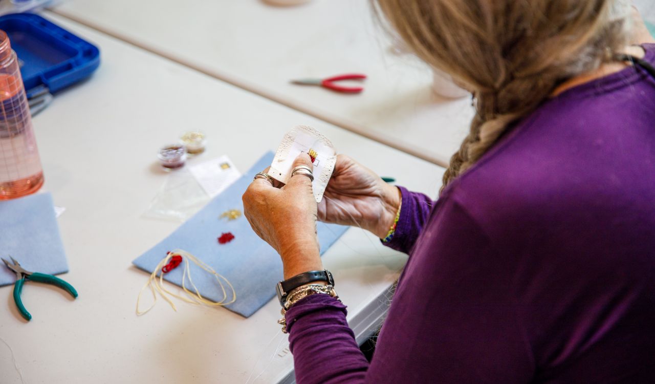 A close-up shot of a woman’s hands shows her beading at a table with tools and materials on the table top.