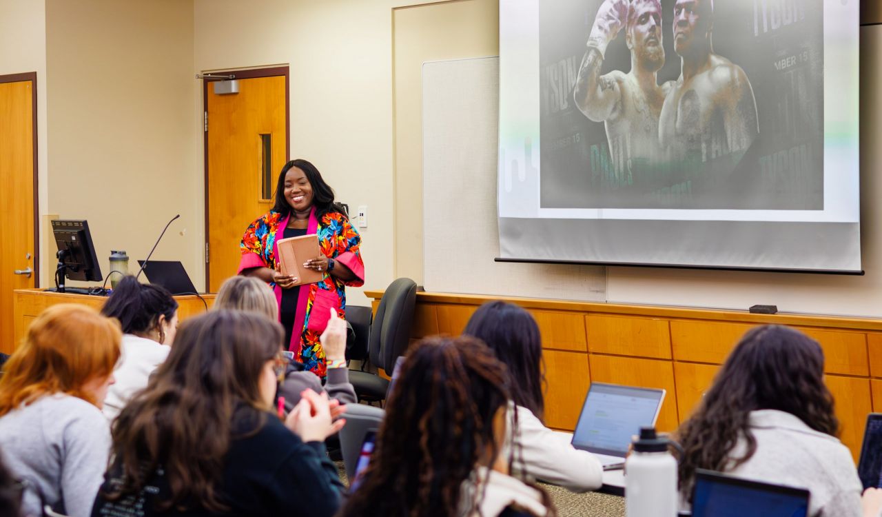 Maame De-Heer, an instructor at Brock University, stands smiling at the front of a classroom holding a notebook. Behind her, a presentation slide features an image of Mike Tyson and Jake Paul under the title 'I.D.E.A. on the Boxing Fight.' Students seated at desks are engaged, with some raising their hands and others working on laptops.