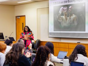 Maame De-Heer, an instructor at Brock University, stands smiling at the front of a classroom holding a notebook. Behind her, a presentation slide features an image of Mike Tyson and Jake Paul under the title 'I.D.E.A. on the Boxing Fight.' Students seated at desks are engaged, with some raising their hands and others working on laptops.