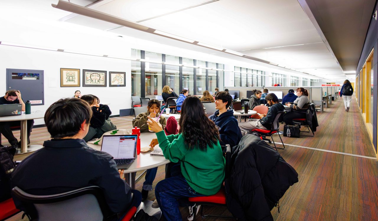 Clusters of students sit in a modern library among tables and study desks surrounded by jackets and backpacks.