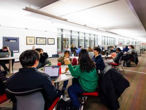 Clusters of students sit in a modern library among tables and study desks surrounded by jackets and backpacks.