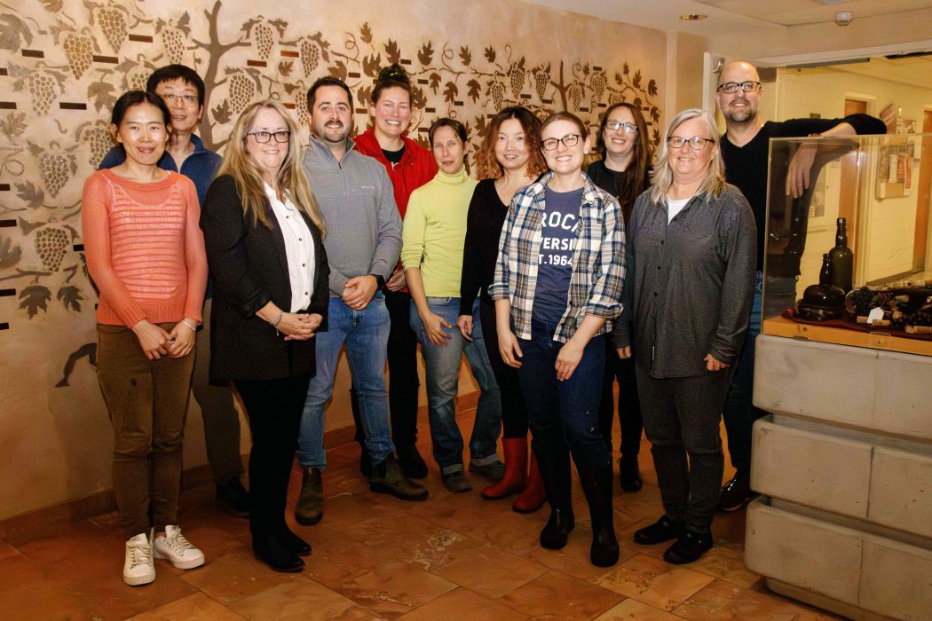 A group of 11 people stand in front of a wall with etchings of grapes and leaves in the background, and a display case on the right containing bottles.