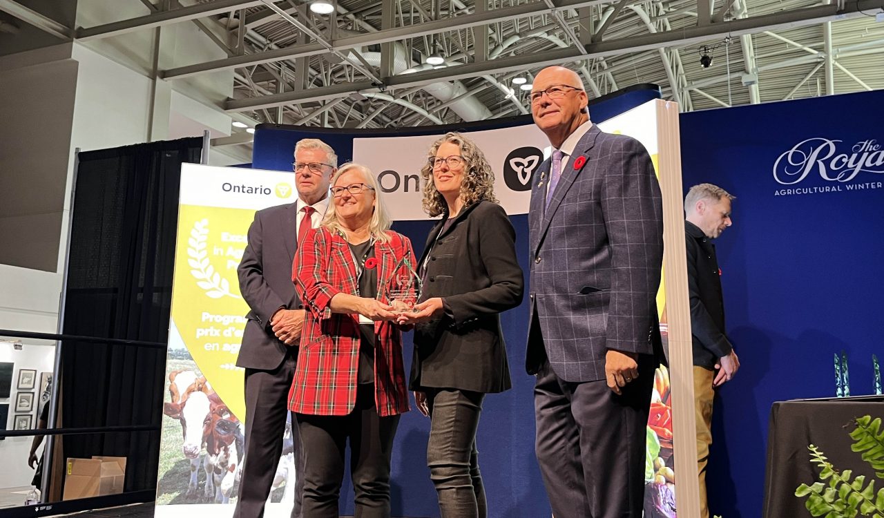 Four people stand in front of two pop-up banners with branding from the Ontario government. The two women in the centre collectively hold a glass award between them.