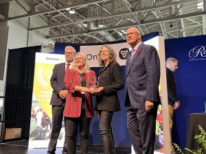 Four people stand in front of two pop-up banners with branding from the Ontario government. The two women in the centre collectively hold a glass award between them.