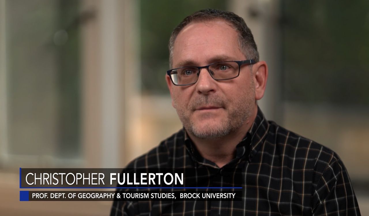 Chris Fullerton wears a plaid, collared shirt and glasses and conducts a television interview while text with his name and title show on screen.